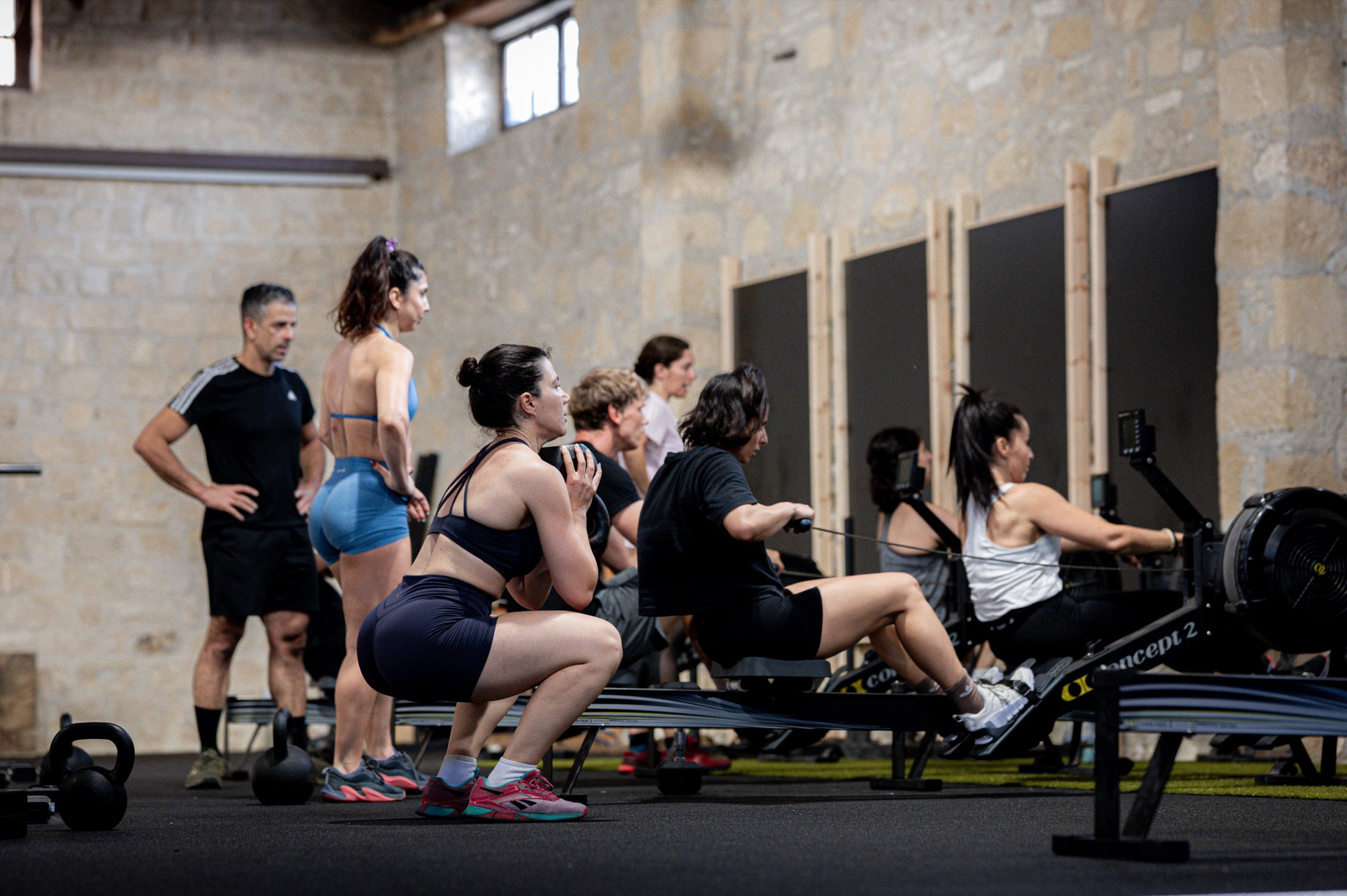 A class on the rowers during a partner workout, with members squatting beside them and a coach watching from the back.