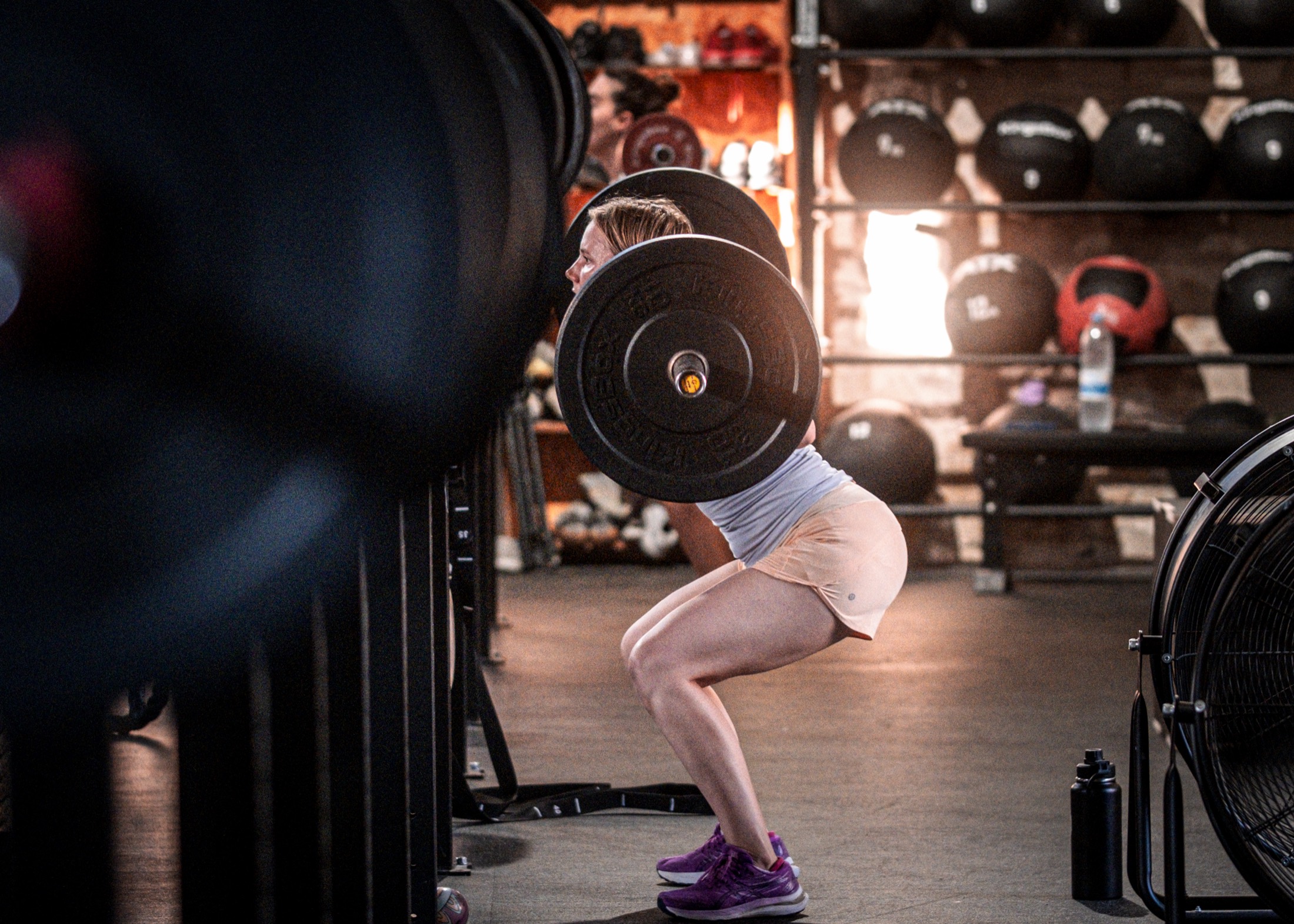 A member in a back squat on the IRONSKY gym floor, medicine balls and plates behind her.