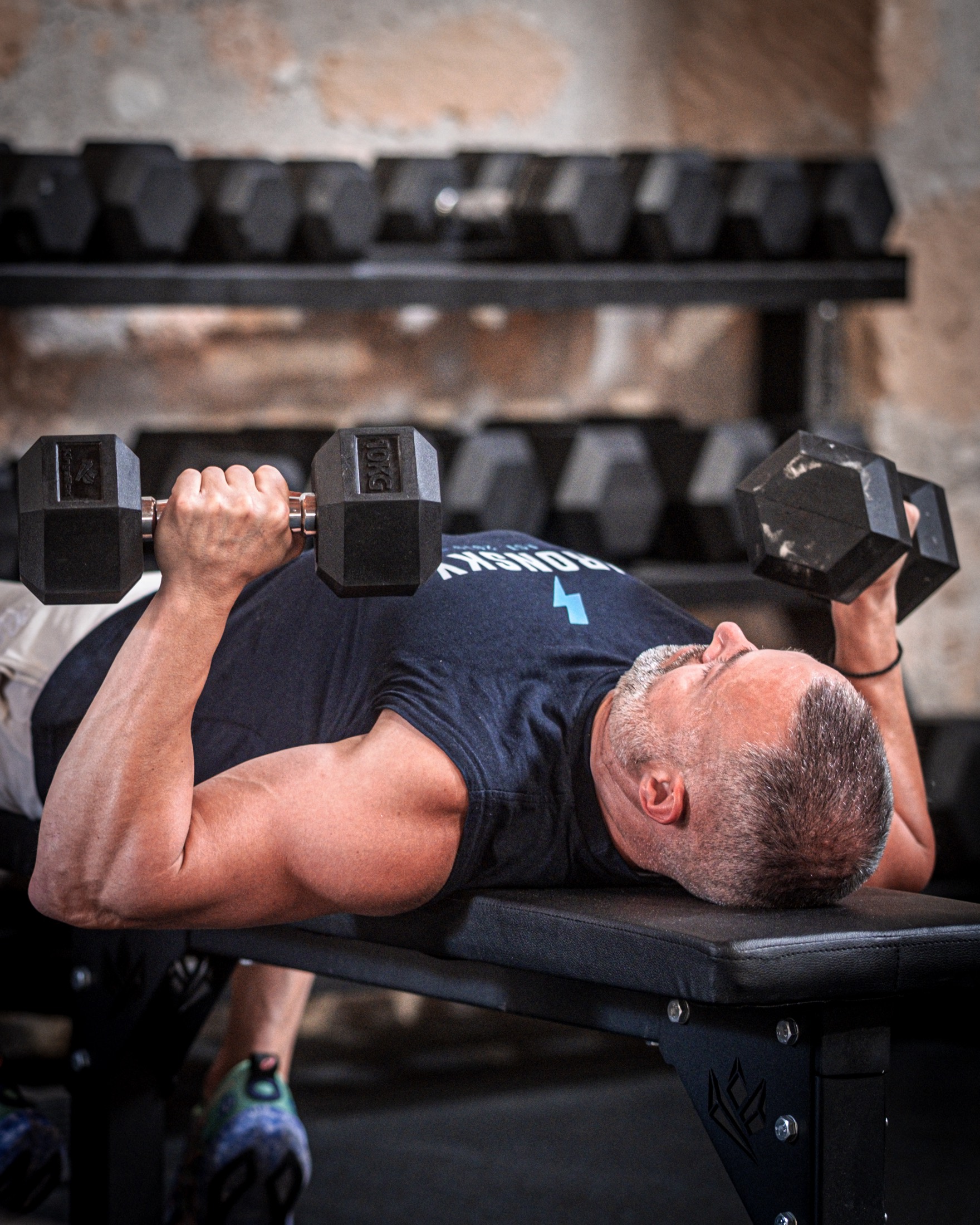 A member in an IRONSKY t-shirt presses two dumbbells on a flat bench in the bodybuilding area, the dumbbell rack behind him.
