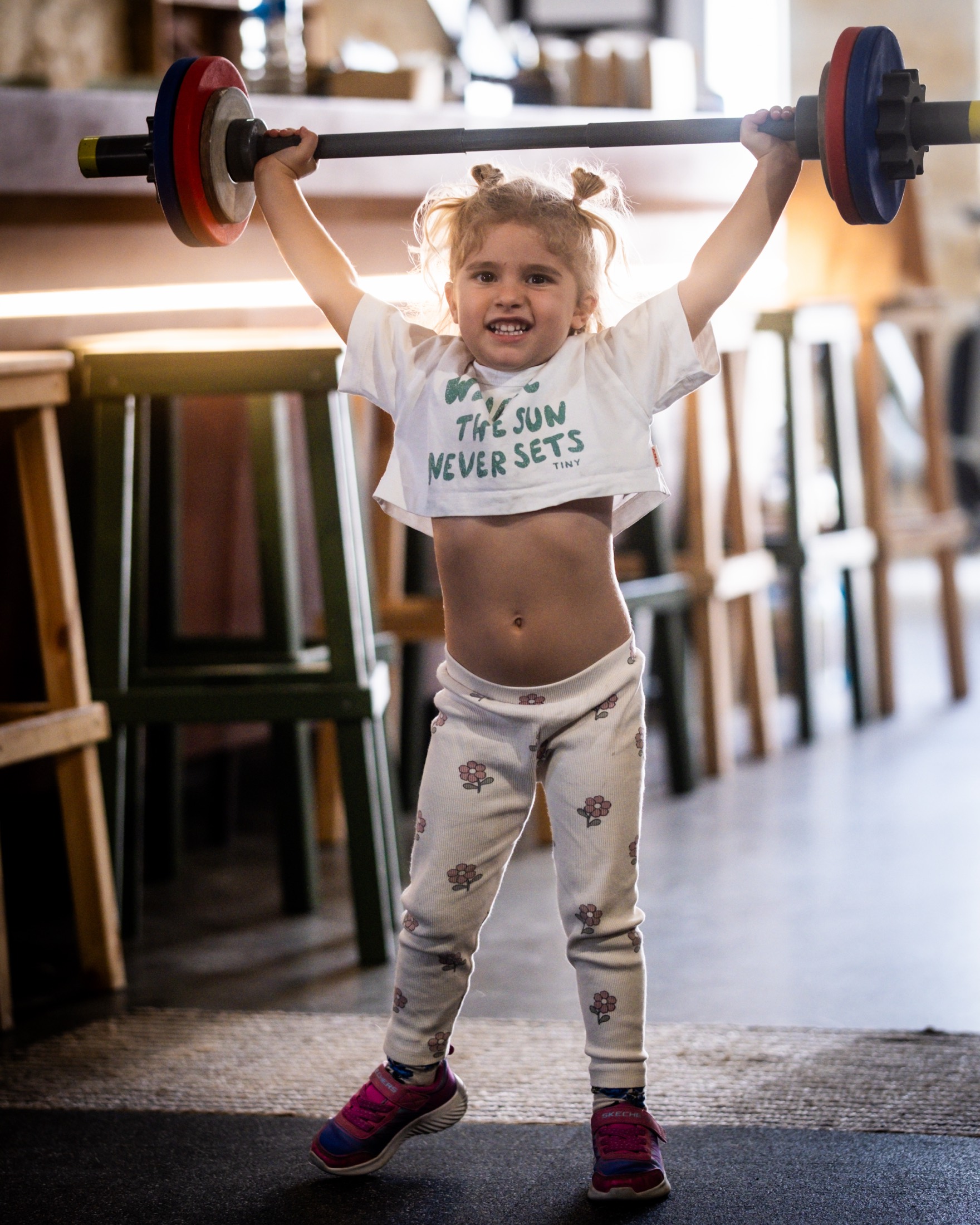 A young girl in the IRONSKY gym holding a light barbell overhead, smiling at the camera.