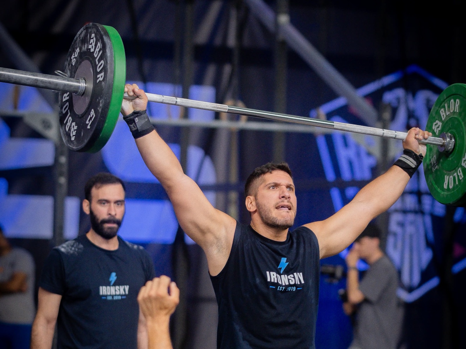 An IRONSKY athlete locks out a loaded barbell overhead on a competition floor while a teammate in IRONSKY kit watches.