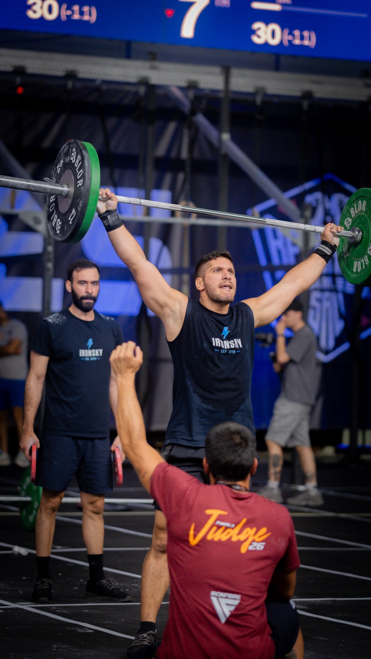 An IRONSKY athlete locks out a loaded barbell overhead at a competition, a teammate watching behind him and a judge in front raising a fist to count the rep.